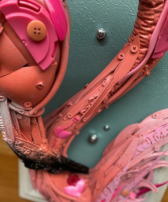 Pink flamingo animal sculpture made from repurposed trash items, displayed in an ornate white frame against a wooden background.