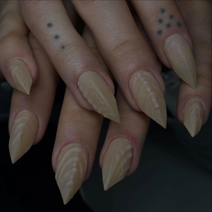 Long n**e almond-shaped nail designs with textured patterns on fingers featuring small dot tattoos, showcasing bold nail designs.