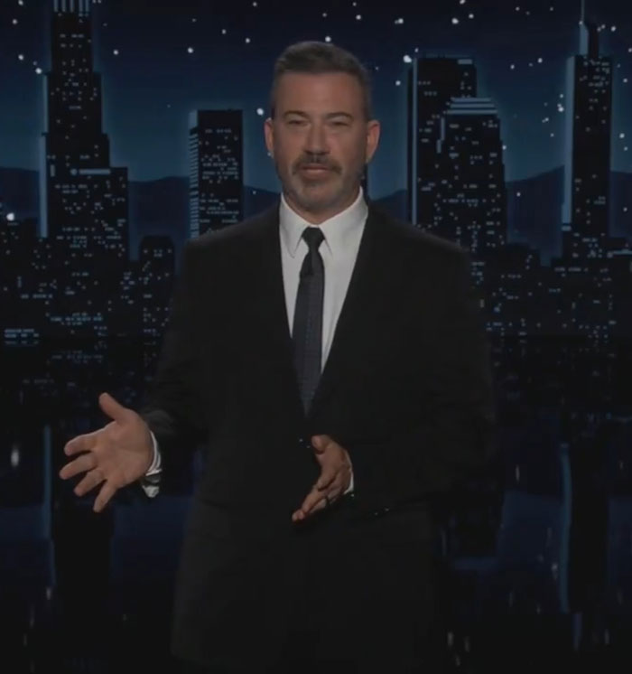 Jimmy Kimmel in a black suit delivering a monologue on a nighttime show set with city skyline background. Jimmy Kimmel in a black suit delivering a monologue on a nighttime show set with city skyline background.