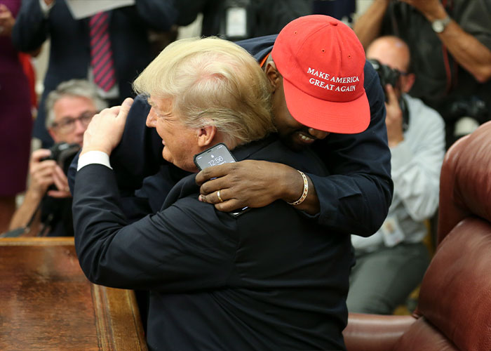 Kanye West wearing red hat hugging former president in a public setting with photographers capturing the moment. Kanye West wearing red hat hugging former president in a public setting with photographers capturing the moment.