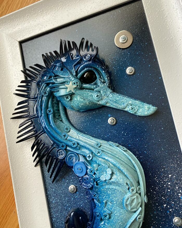 Blue seahorse sculpture made from trash and scrap materials, displayed in a white frame on a wooden floor.