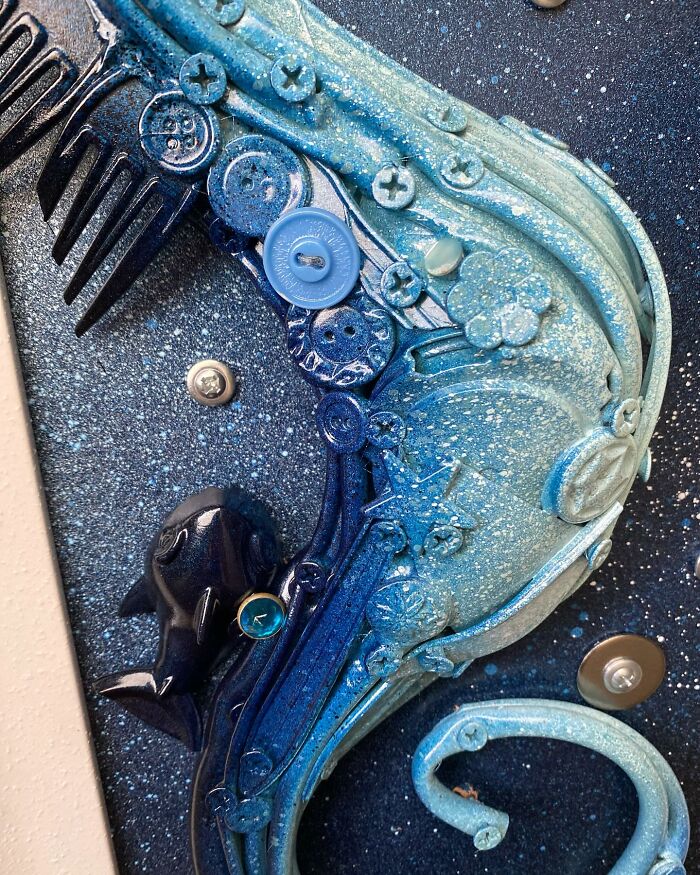 Blue seahorse sculpture made from trash and scrap materials, displayed in a white frame on a wooden floor.