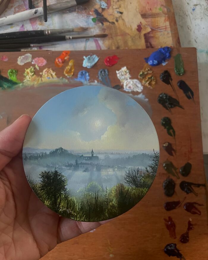 Hand holding a detailed circular painting by Dina Brodsky depicting a misty natural landscape with trees and distant buildings.