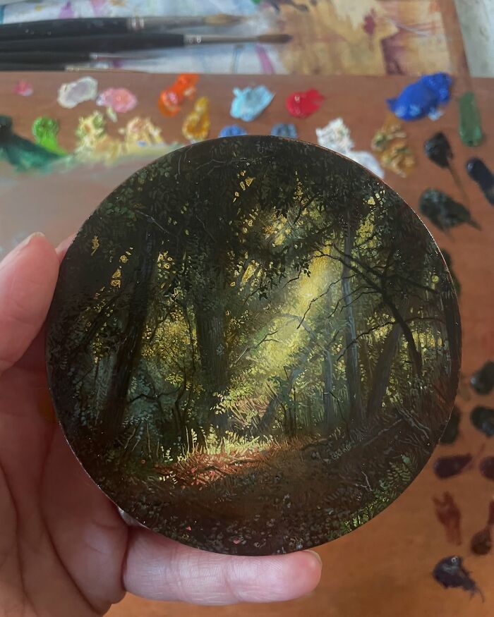 Hand holding a detailed painting by Dina Brodsky depicting a sunlit forest path and the beauty of the natural world.