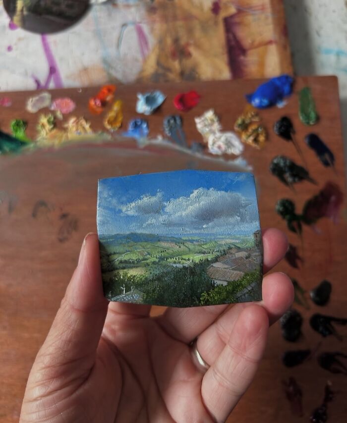 Hand holding a detailed painting of a natural landscape with blue sky, green hills, and clouds by Dina Brodsky.