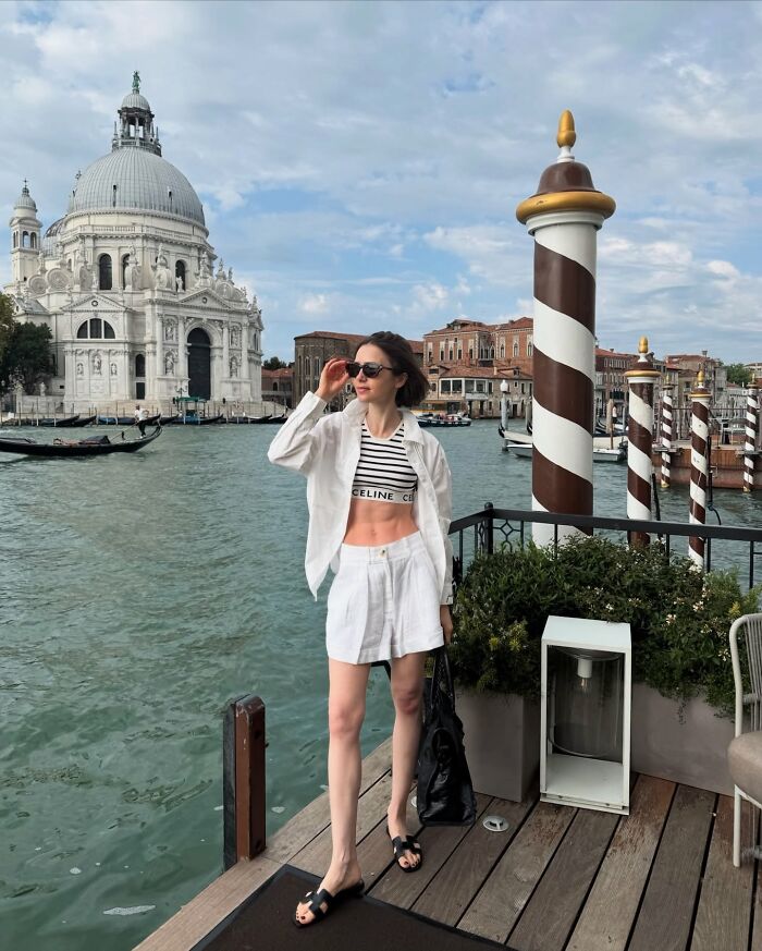 Lily Collins with extreme thinness, wearing white outfit and sunglasses, standing by Venice canal and historic architecture. Lily Collins with extreme thinness, wearing white outfit and sunglasses, standing by Venice canal and historic architecture.