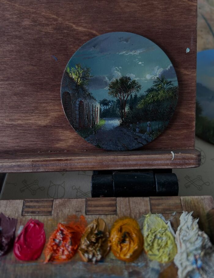 Round detailed painting of a natural landscape with trees and a path, surrounded by vibrant paint colors on a wooden surface.