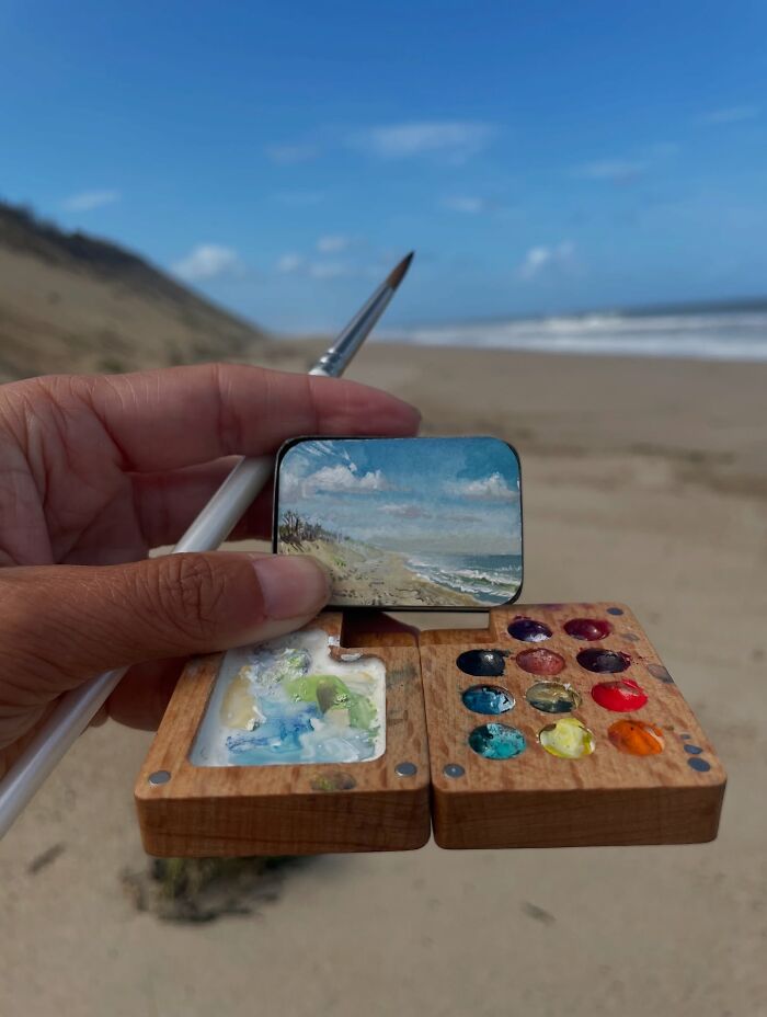 Hand holding a miniature coastal painting and paintbrush with a wooden palette, showcasing detailed natural world art.
