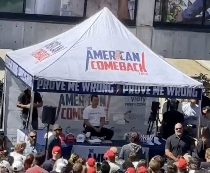 Crowd gathered near commentator booth with The American Comeback tent and people wearing red hats at outdoor event. Crowd gathered near commentator booth with The American Comeback tent and people wearing red hats at outdoor event.
