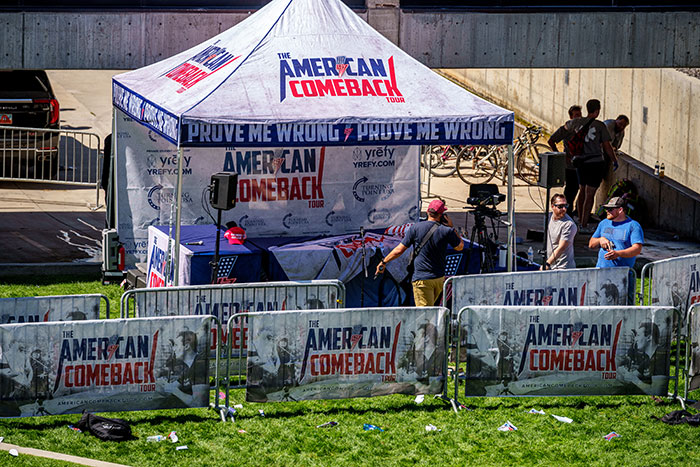 Outdoor tent setup with American Comeback Tour banners and people setting up recording equipment near metal barricades. Outdoor tent setup with American Comeback Tour banners and people setting up recording equipment near metal barricades.