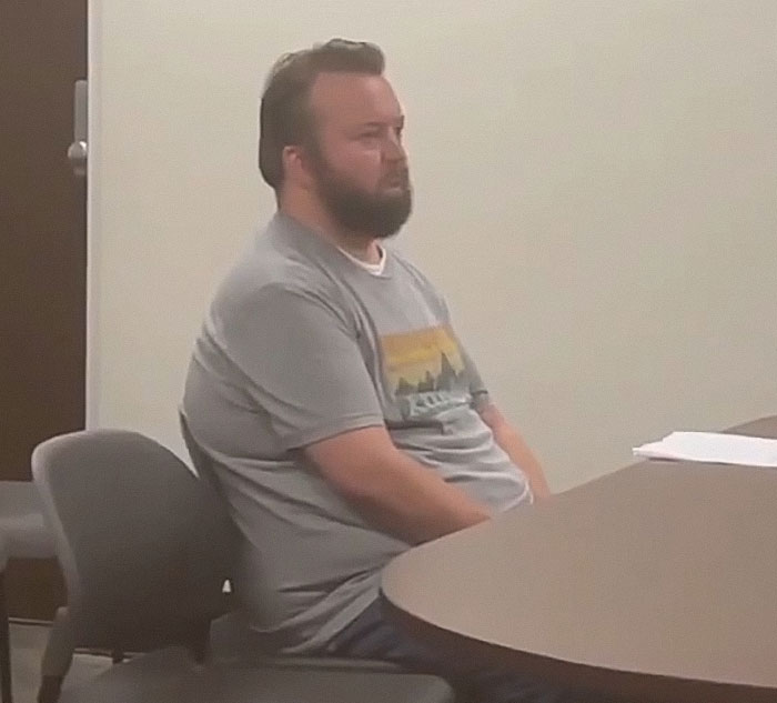 Man with beard sitting at a table in a plain room, related to teacher arrested for using poop spray at school causing damage. Man with beard sitting at a table in a plain room, related to teacher arrested for using poop spray at school causing damage.