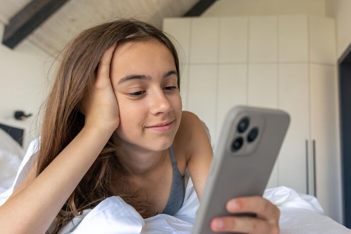 Young woman lying on bed, looking at her smartphone with a slight smile, reflecting childish reasons for ditching a guy.