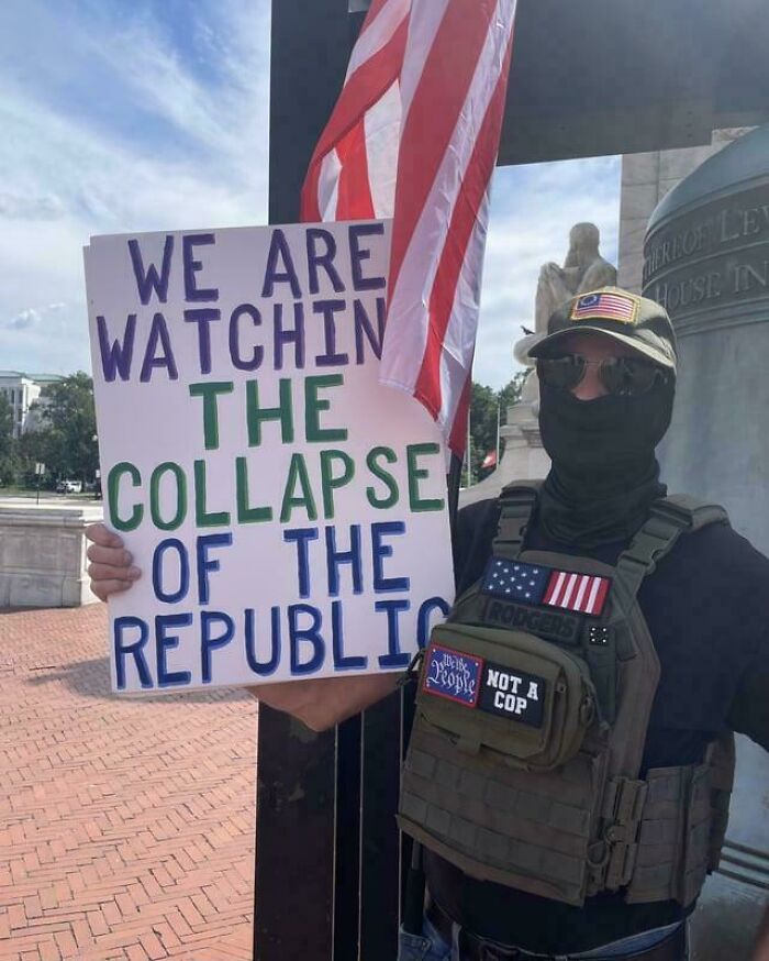 Person wearing tactical gear and holding a protest sign about the collapse of the republic, embodying chaotic good actions.