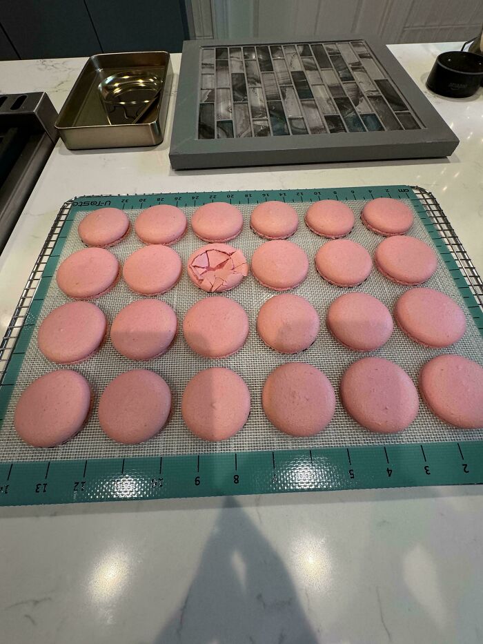 Pink macarons cooling on a mat, with one c*****d piece disrupting the neat rows, showing cute cat paw prints humor concept.