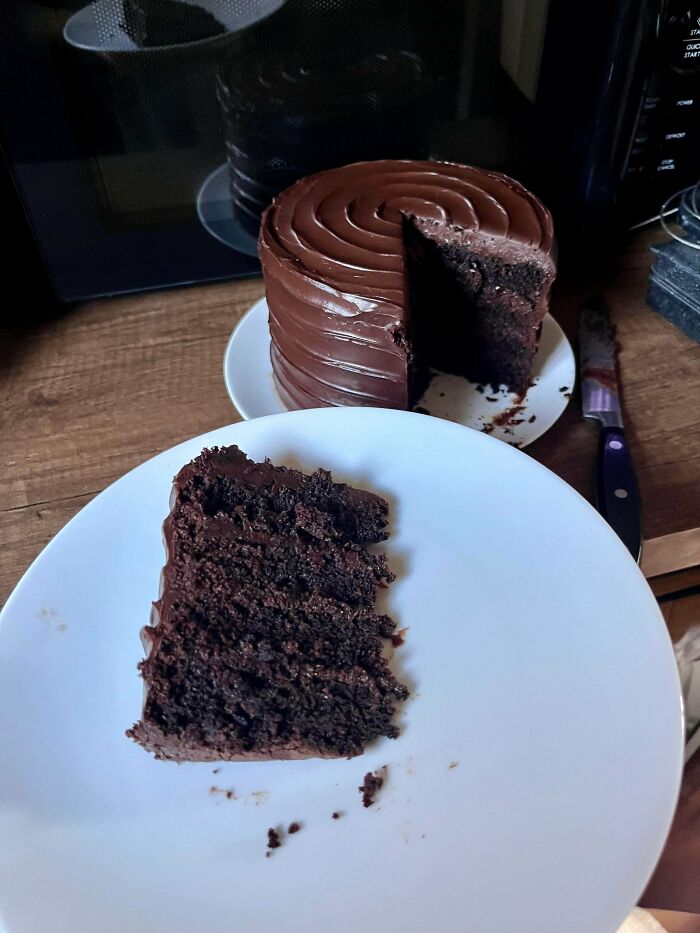 Slice of rich chocolate cake with smooth frosting on a white plate beside the rest of the baked goods on a wooden surface.
