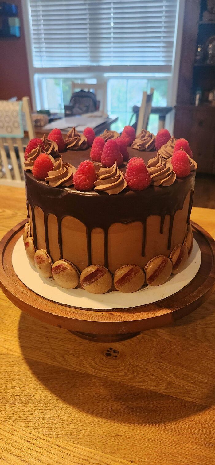Chocolate drip cake topped with fresh raspberries, chocolate frosting swirls, and decorated with macarons on a wooden cake stand.