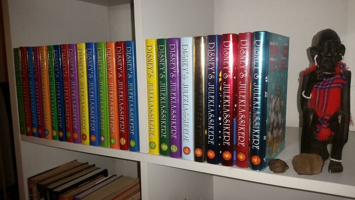 Colorful Danish book series neatly arranged on a white shelf beside a traditional wooden Danish figure and small stones.