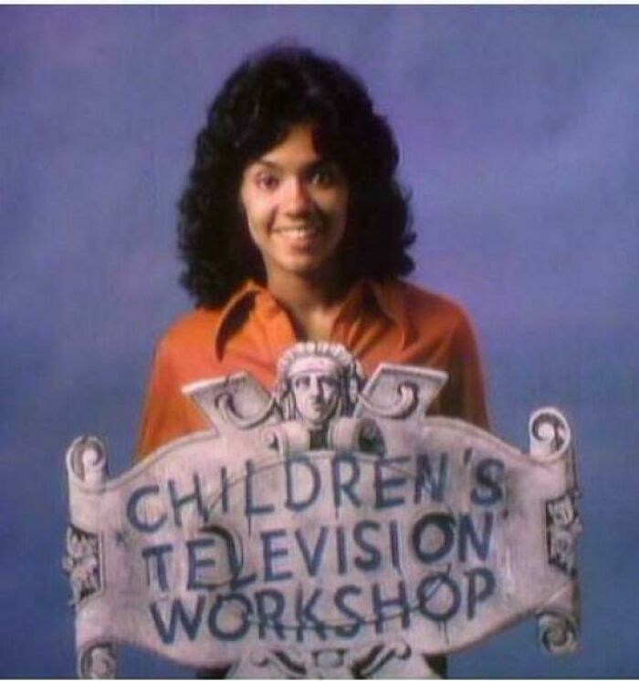 Person with curly hair smiling and holding a vintage Children's Television Workshop sign to make you feel old as heck.