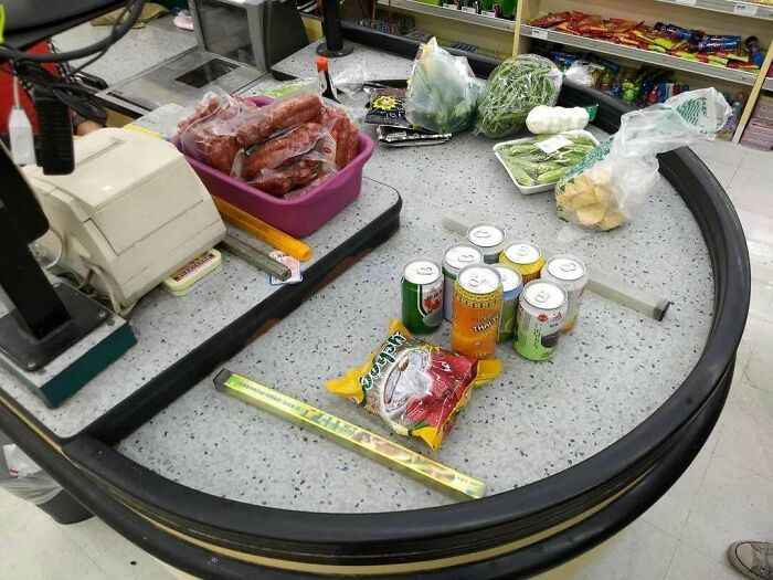 Grocery checkout counter with old-style cash register, canned drinks, packaged sausages, and fresh vegetables visible.