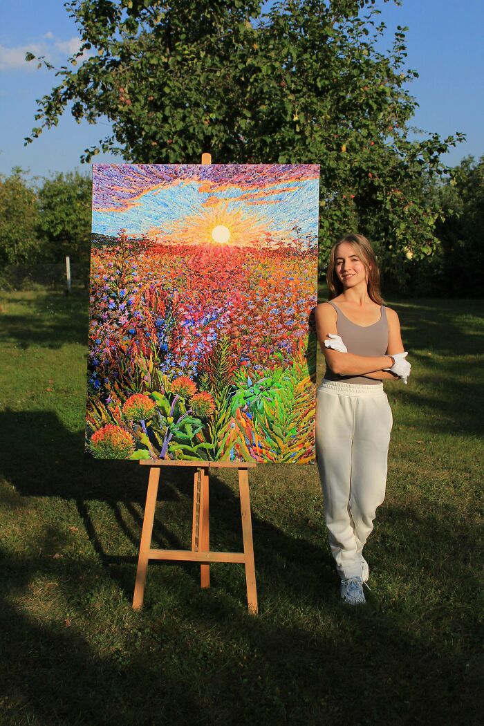 Young woman standing next to a colorful painting of a vibrant sunset over a field, showcasing cool creative art made outdoors.
