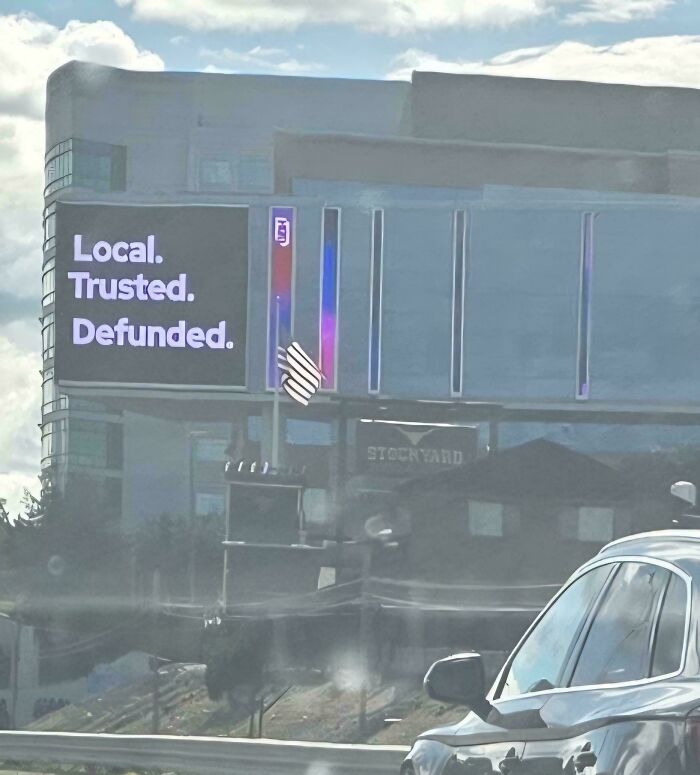 Billboard with text Local Trusted Defunded near highway and modern building in a dystopian nightmare scene in the US.