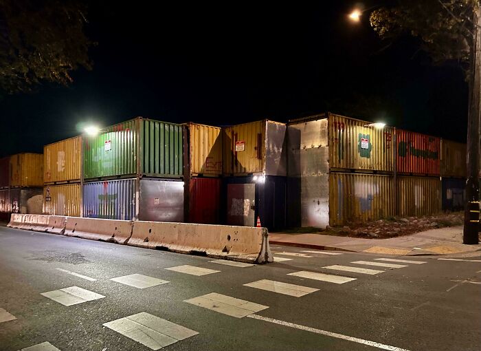 Stacked shipping containers and concrete barriers blocking a street at night, resembling a dystopian scene in the US.