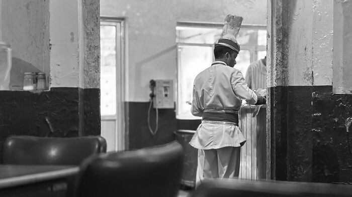 Black and white street photography capturing a man in traditional attire inside a modest room, highlighting raw humanity.