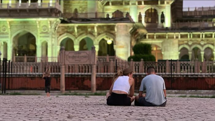 Couple sitting on cobblestone street with historic building in background, a moment of street photography capturing humanity.