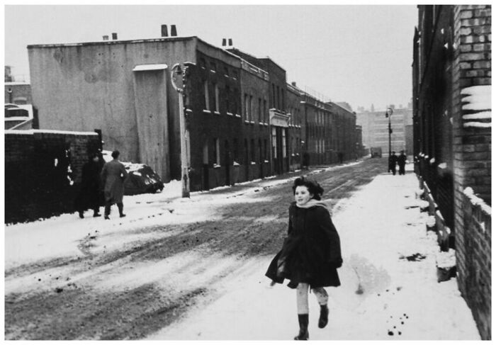 Black and white old photo showing a child running on a snowy street in a residential area, capturing life from a different time.