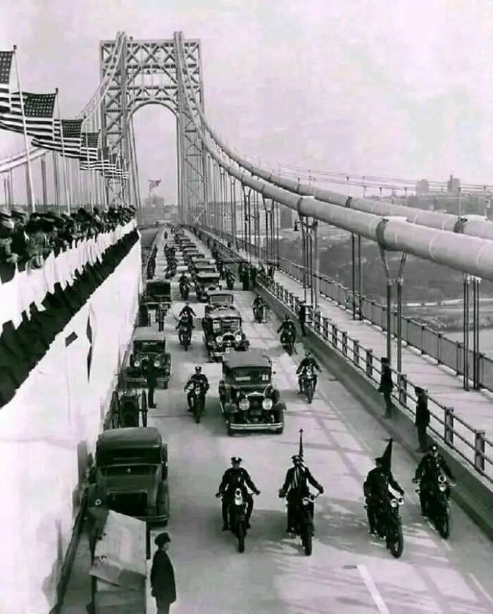 Black and white vintage photo showing old cars and motorcycles on a bridge with American flags and onlookers from a different time.