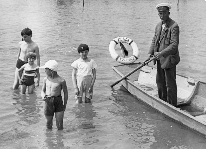 Children wading in water near a man in a boat, captured in an old photo showing people’s lives from a different time.
