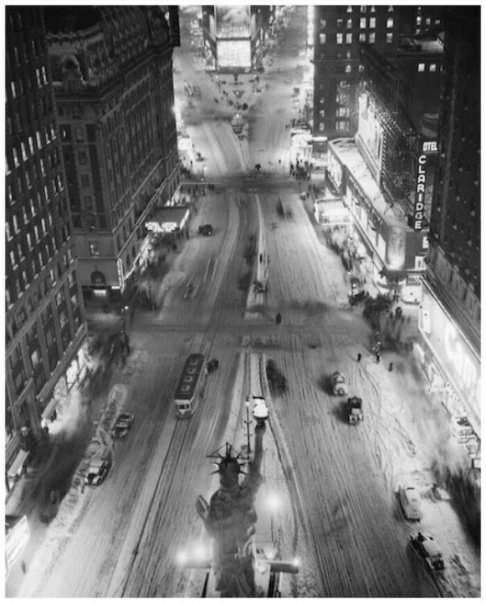 Black and white old photo showing a snowy city street with cars and pedestrians from a different time period.