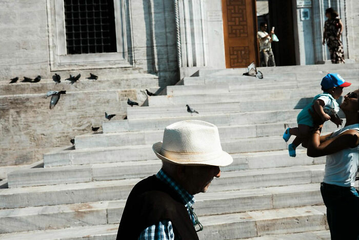 An elderly man wearing a hat in front of stone steps with pigeons and a father lifting his child in Turkey.