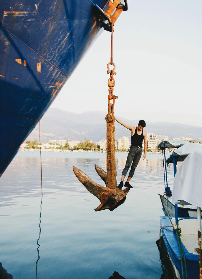 Man balancing on a large anchor hanging from a ship, capturing candid life moments in Turkey's coastal harbor.
