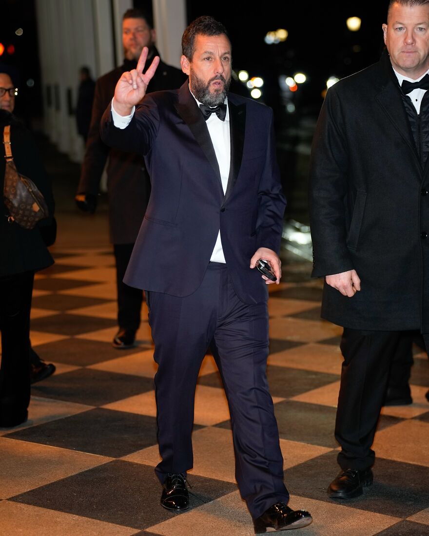 Adam Sandler wearing a navy tuxedo and bow tie, making a peace sign while walking on a checkered floor at night.