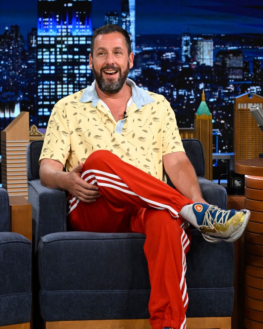 Adam Sandler in a retro yellow patterned shirt and red track pants on a late-night talk show set.