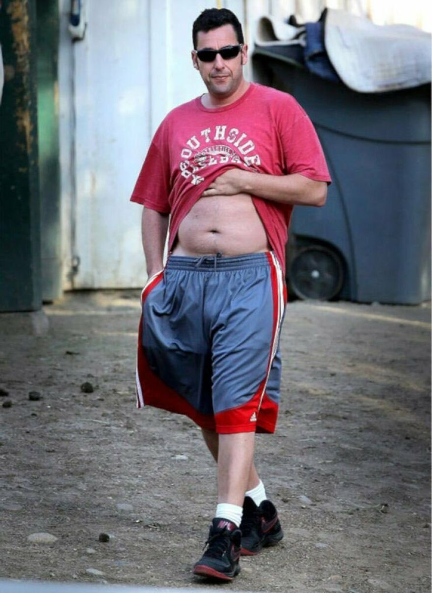 Adam Sandler in casual outfit with red t-shirt lifted, gray and red shorts, black sneakers, and sunglasses outdoors.