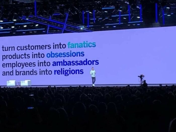 Speaker on stage in front of a large audience with a presentation about fanatics, obsessions, ambassadors, and religions.