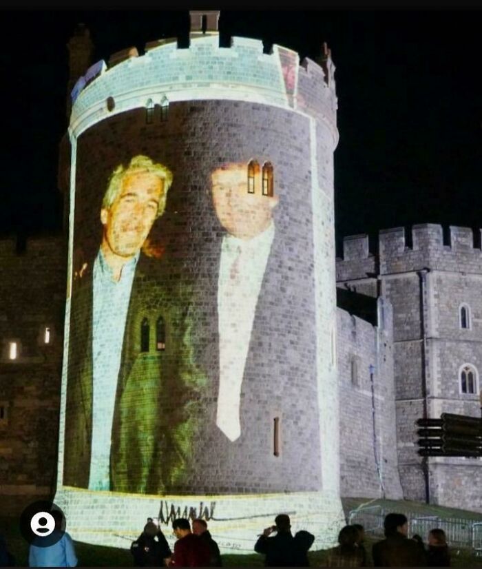 Projection of a large image on a castle tower, showing two men, with people watching below at night.