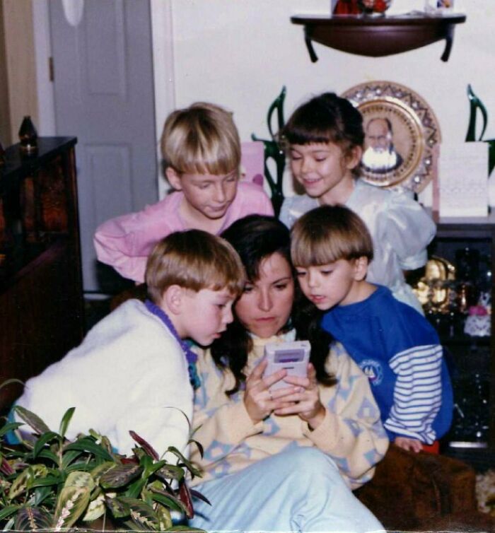 Mom and children gathered closely while she uses a handheld game console, capturing a wholesome family moment.