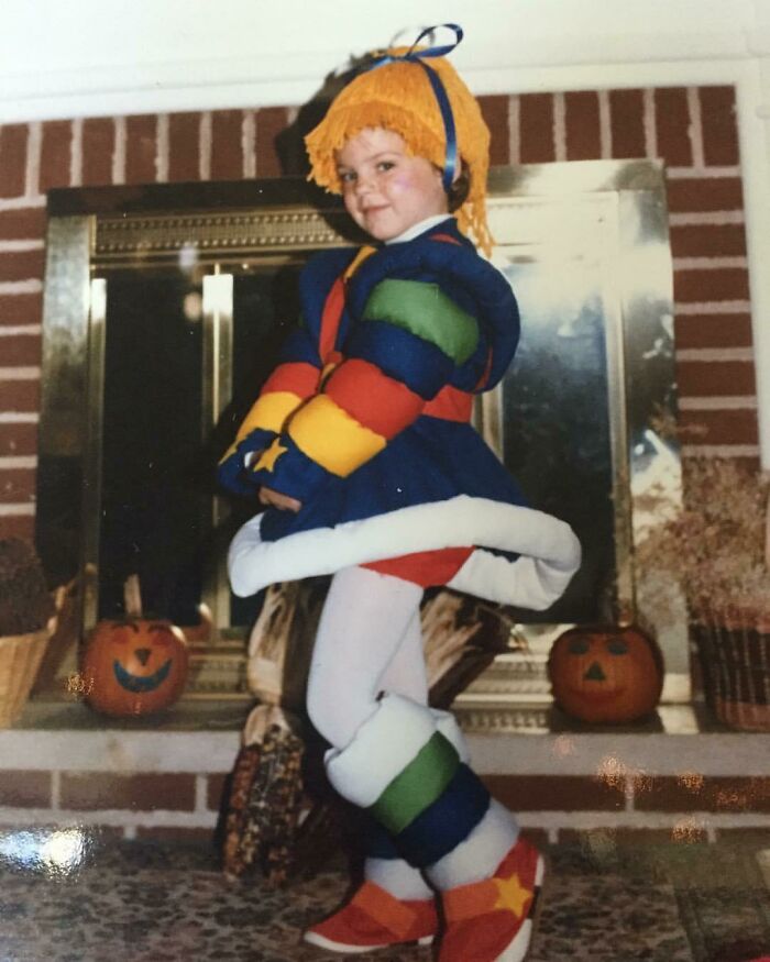 Child dressed in colorful costume standing indoors near festive pumpkin decorations, capturing wholesome mom moments.