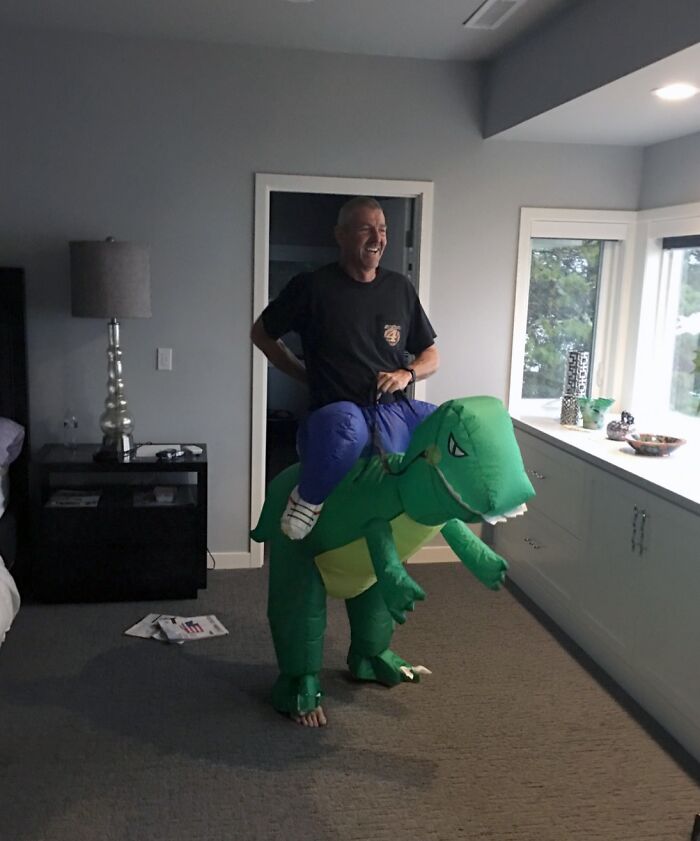 Man wearing inflatable dinosaur costume indoors, laughing and having fun, showing one of the funniest parents ever moments.
