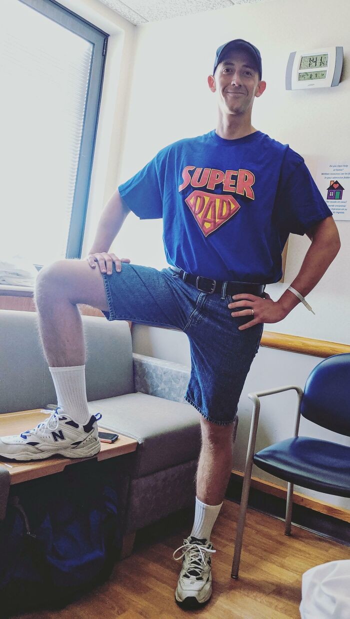Man wearing a Super Dad shirt and denim shorts posing with one leg on a table in a room, funny parents vibe.