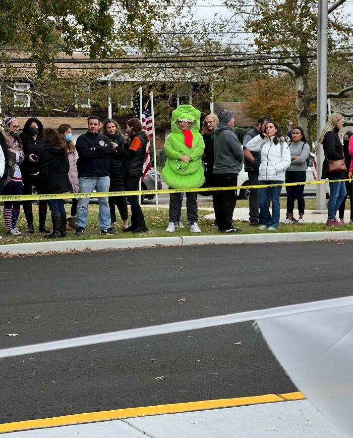 Group of people standing behind caution tape outdoors, including one person dressed in a bright green dinosaur costume, showing funniest parents vibe.