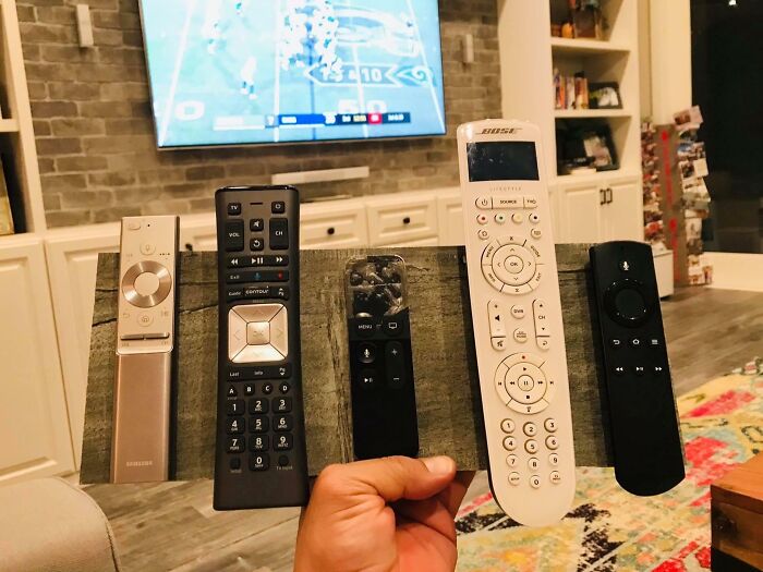 Hand holding a board with five TV remotes attached, showing a funny parents' creative solution in a living room setting.