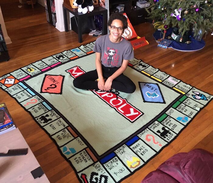 Person sitting on a large Monopoly-themed rug in a living room with a Christmas tree and hardwood floor.