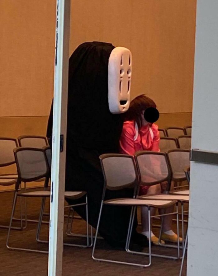 Person dressed as No Face character sitting next to child in a pink outfit, a funny parents moment captured indoors.