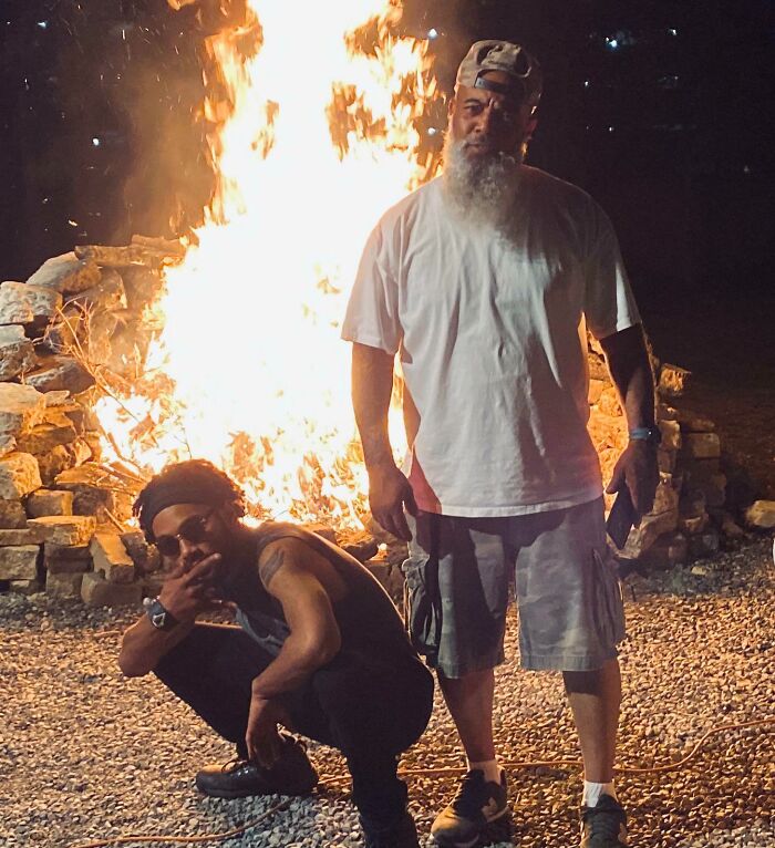 Man with a white beard and cap stands by fire pit while younger man crouches nearby in a funny parents moment outdoors.