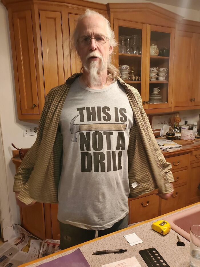 Older man in a kitchen wearing a humorous shirt that says This is Not a Drill, showcasing funny parents moments.