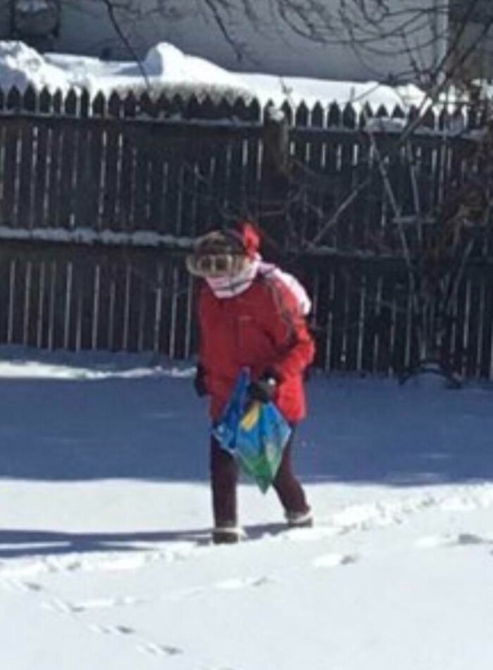 Mom in a red coat walking through snow in a backyard, carrying a colorful bag on a bright winter day.